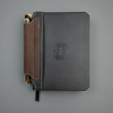 Load image into Gallery viewer, Franklin-Christoph black and brown leather elastic pen holder with creme stitching on a black notebook.